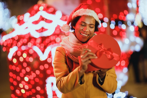 Sorridente donna nera che tiene un regalo sul mercatino di Natale festivo all'aperto nella notte d'inverno in città.