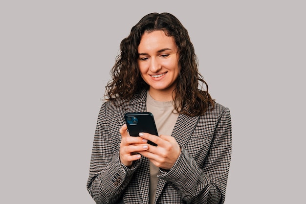 Sorridente donna riccia sta scrivendo un messaggio o navigando in internet al telefono