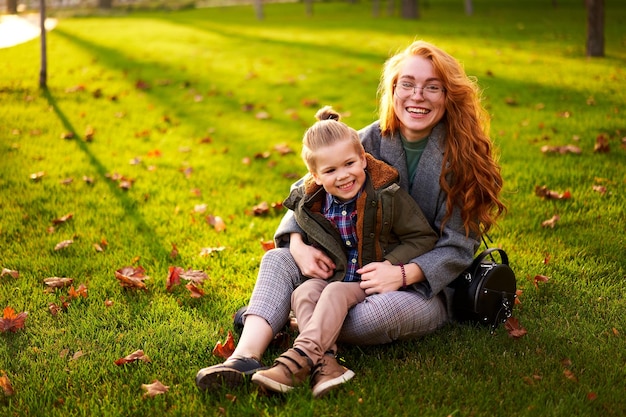 Sorridente donna rossa e ragazzino seduto sul prato nel parco cittadino il caldo giorno d'autunno giovane mamma