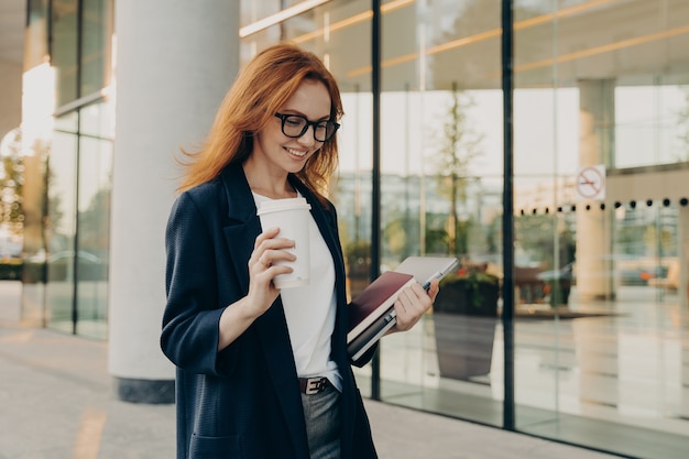 Sorridente donna rossa lavoratrice aziendale vestita con abiti formali beve caffè