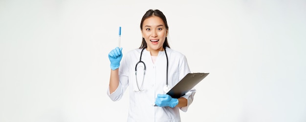 Sorridente dottoressa asiatica alzando la penna eureka gesto che tiene appunti in piedi in uniforme medica su sfondo bianco