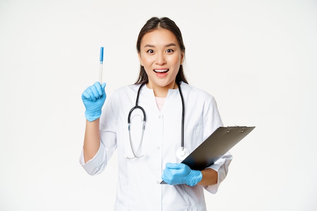 Sorridente dottoressa asiatica alzando la penna, gesto di eureka, tenendo la lavagna per appunti, in piedi in uniforme medica su sfondo bianco.