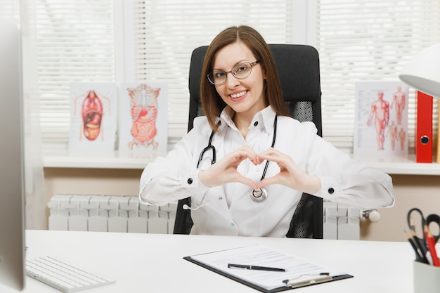 Sorridente dottoressa seduta alla scrivania, che mostra la forma del cuore con le mani, lavorando con documenti medici in ufficio leggero in ospedale