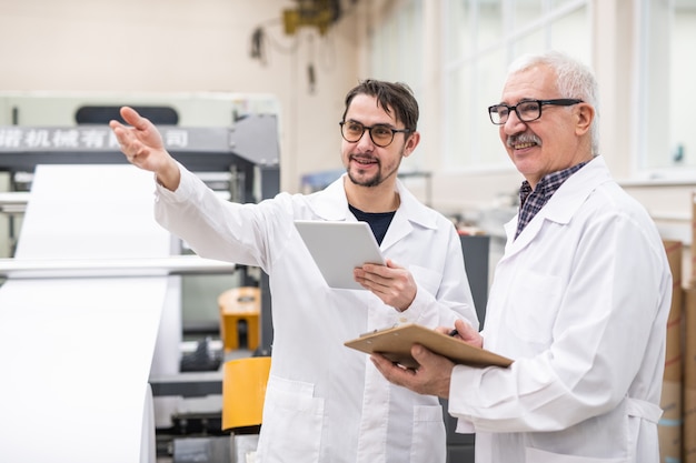 Sorridente esperto di qualità barbuto in bicchieri gesticolando mano e utilizzo di tablet mentre discute la produttività delle attrezzature per la stampa con il collega