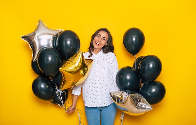 Sorridente felice ragazza carina eccitata che tiene in mano molti palloncini neri sta posando sullo sfondo giallo mentre fa shopping il Black Friday