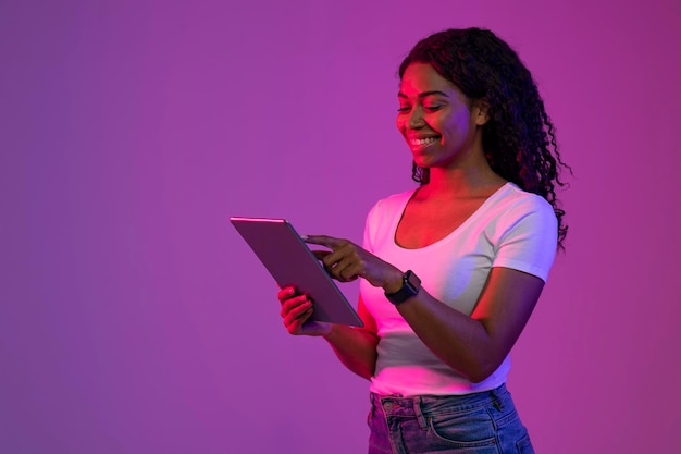 Sorridente femmina nera utilizzando la tavoletta digitale mentre si sta in piedi nella luce al neon