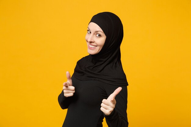 Sorridente fiduciosa bella giovane donna musulmana araba in abiti neri hijab in posa isolata sulla parete gialla, ritratto. Concetto di stile di vita dell'Islam religioso della gente.