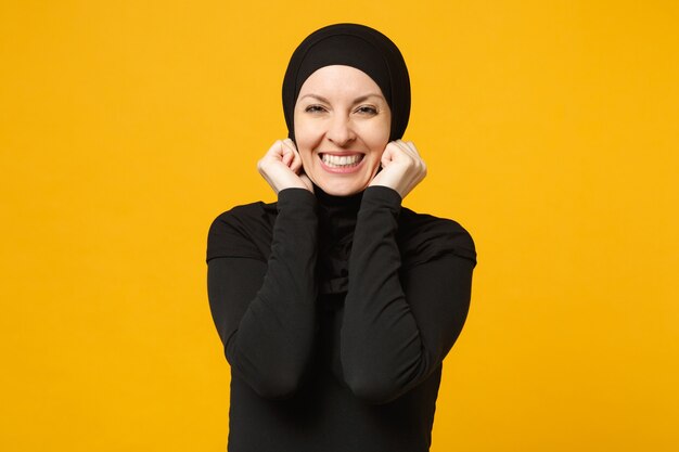 Sorridente fiduciosa bella giovane donna musulmana araba in abiti neri hijab in posa isolata sulla parete gialla, ritratto. Concetto di stile di vita dell'Islam religioso della gente.