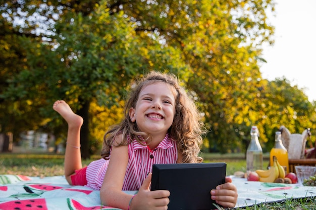 Sorridente fiducioso bambino sdraiato sul tappetino da picnic tiene il suo tablet