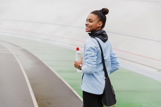 Sorridente fit giovane sportiva africana che trasporta borsa sportiva in piedi allo stadio, tenendo in mano una bottiglia d'acqua