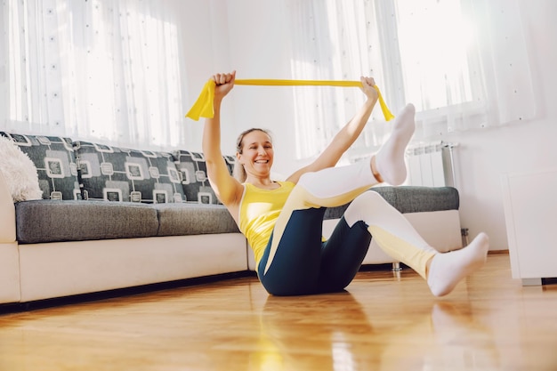 Sorridente forte sportiva in forma seduta sul pavimento a casa e allungando la gomma di potenza con le mani