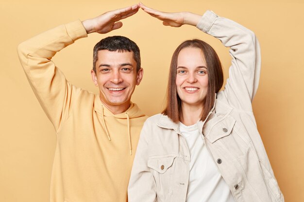 Sorridente gioiosa allegra giovane uomo e donna coppia in piedi insieme isolato su sfondo beige facendo tetto con le mani vuole la propria casa