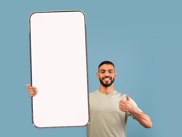 Sorridente giovane arabo che dimostra smartphone gigante con mockup che mostra i pollici in su per la promozione