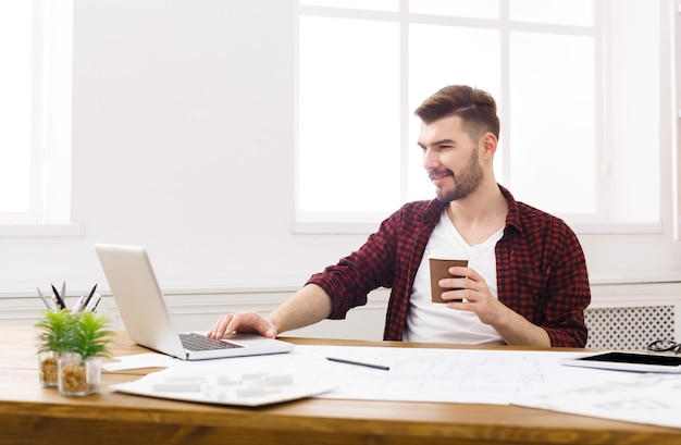 Sorridente giovane architetto che lavora casualmente con progetti e laptop in interni moderni sul posto di lavoro, copia spazio.