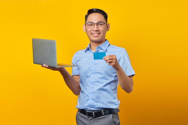 Sorridente giovane asiatico asiatico con gli occhiali in possesso di laptop e mostrando la carta di credito isolata su sfondo giallo concetto di uomo d'affari e imprenditore