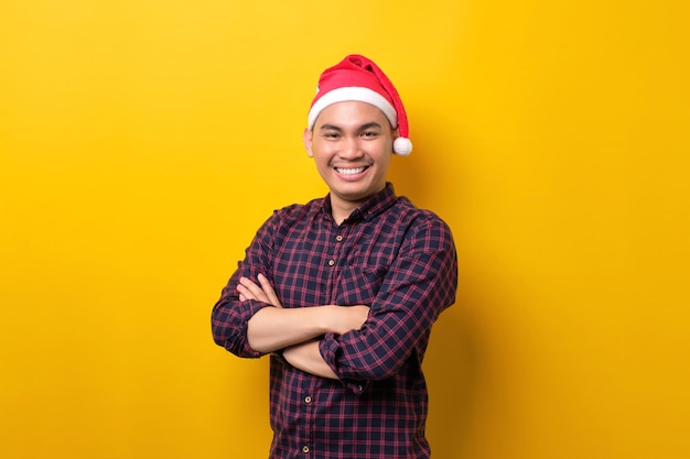 Sorridente giovane asiatico con cappello da Babbo Natale in piedi con le braccia incrociate guardando la fotocamera su sfondo giallo studio Happy New Year 2023 celebrazione buon concetto di vacanza