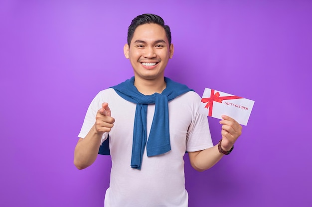 Sorridente giovane asiatico in abiti casual tenendo buono regalo e puntando il dito la fotocamera isolata su sfondo viola studio
