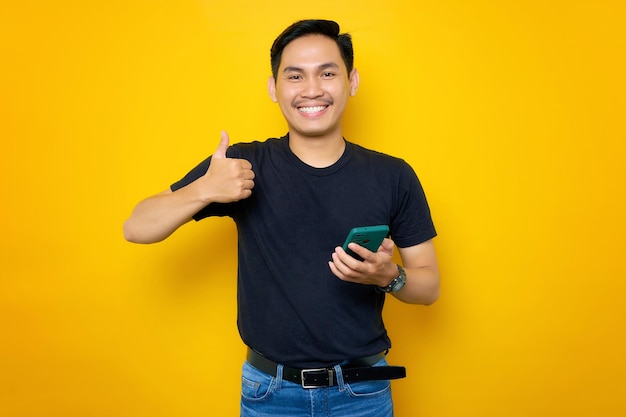 Sorridente giovane asiatico in maglietta casual che tiene smartphone e mostra il gesto del pollice in su isolato su sfondo giallo Concetto di stile di vita delle persone