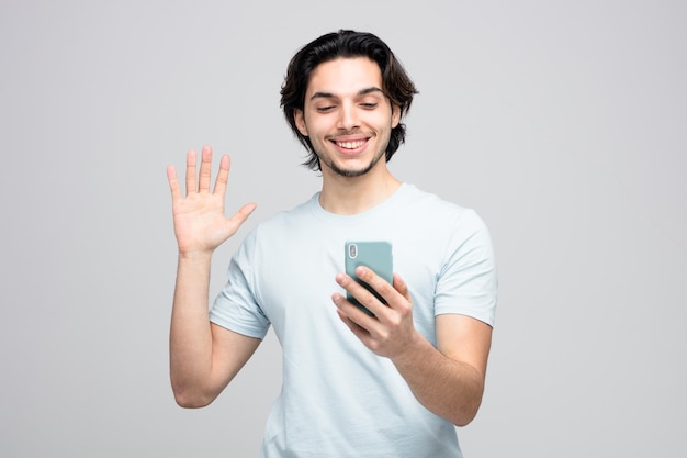 sorridente giovane bell'uomo che tiene il telefono cellulare parlando tramite videochiamata sventolando isolato su sfondo bianco