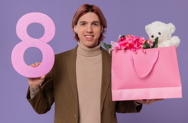 Sorridente giovane bell'uomo con numero rosa otto e sacchetto regalo con fiori e orsacchiotto