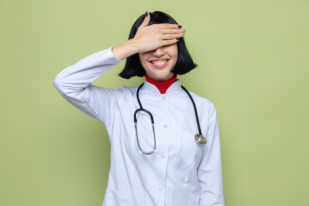 Sorridente giovane bella donna caucasica in uniforme da medico con stetoscopio che copre gli occhi con la mano