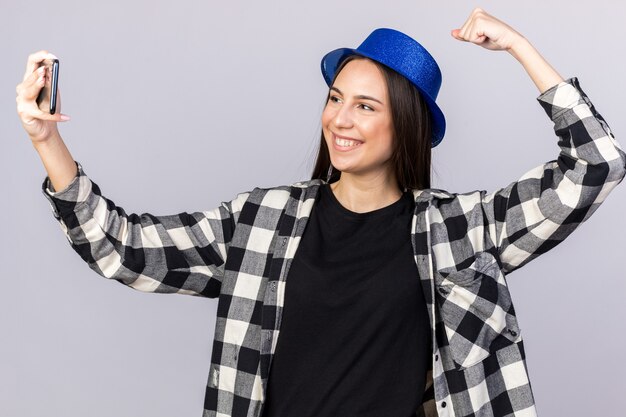 Sorridente giovane bella donna che indossa un cappello da festa che fa un gesto forte, scatta un selfie isolato sul muro bianco