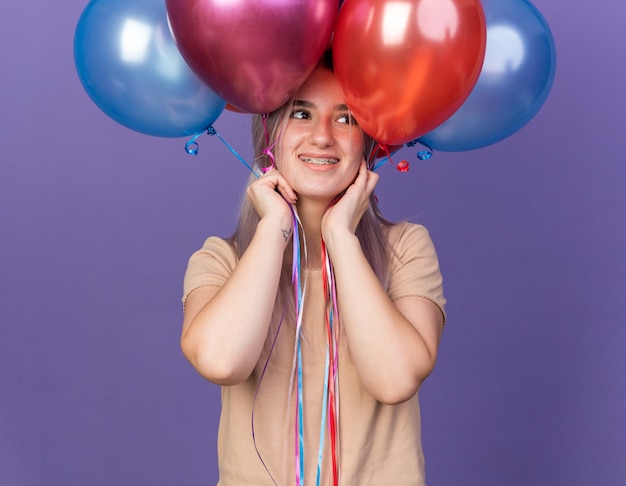 Sorridente giovane bella ragazza che indossa bretelle dentali che tengono palloncini sulla testa isolata sulla parete blu