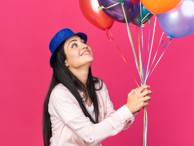 Sorridente giovane bella ragazza che indossa cappello da festa tenendo e guardando i palloncini