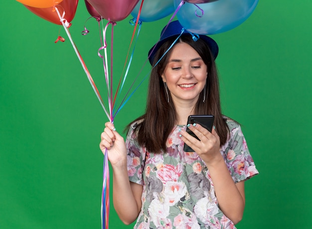 Sorridente giovane bella ragazza che indossa un cappello da festa con palloncini e guardando il telefono in mano isolato sul muro verde