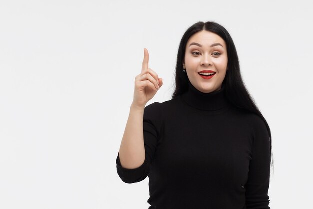 Sorridente giovane bruna stupita in dolcevita nero che ha idea e punta l'indice verso l'alto