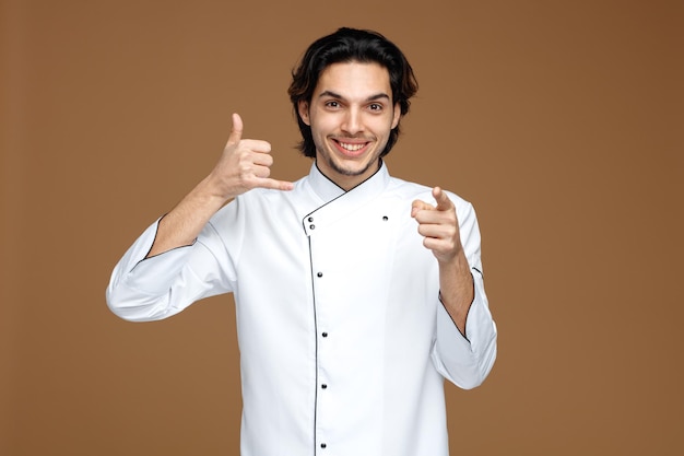 sorridente giovane chef maschio che indossa l'uniforme guardando e indicando la fotocamera che mostra il gesto di chiamata isolato su sfondo marrone