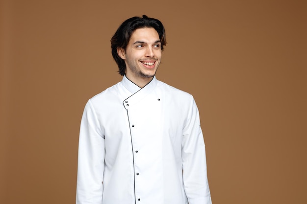 sorridente giovane chef maschio che indossa uniforme guardando il lato isolato su sfondo marrone