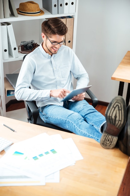 Sorridente giovane con le gambe sul tavolo utilizzando tablet in office