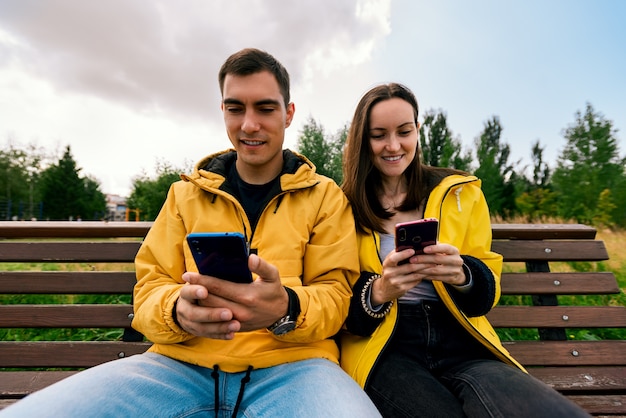 Sorridente giovane coppia in giacche gialle, seduta su una panchina nel parco in autunno utilizzando telefoni, smartphone. Millennials, inseparabili da gadget, dispositivo