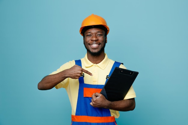 Sorridente giovane costruttore afroamericano in tenuta uniforme e punti negli appunti isolati su sfondo blu