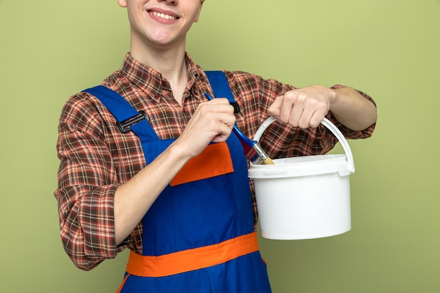 Sorridente giovane costruttore maschio che indossa l'uniforme che tiene il secchio con il pennello