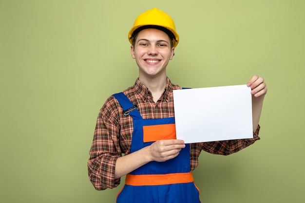 Sorridente giovane costruttore maschio tenendo la carta indossando l'uniforme