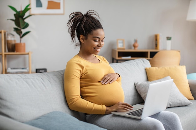 Sorridente giovane donna afroamericana incinta con grande pancia che lavora al computer portatile nell'interno del soggiorno