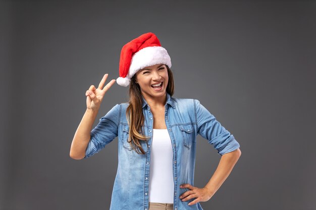 Sorridente giovane donna allegra con il cappello di Babbo Natale sulla testa vestito casual tenendo premuto il segno di vittoria.