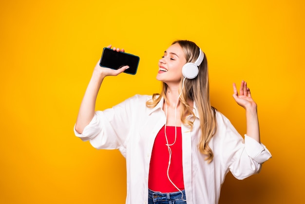 Sorridente giovane donna ascoltando musica in cuffia e utilizzando smartphone isolato sopra la parete gialla