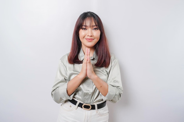 Sorridente giovane donna asiatica che indossa camicia verde salvia gesturing saluto tradizionale isolato su sfondo bianco