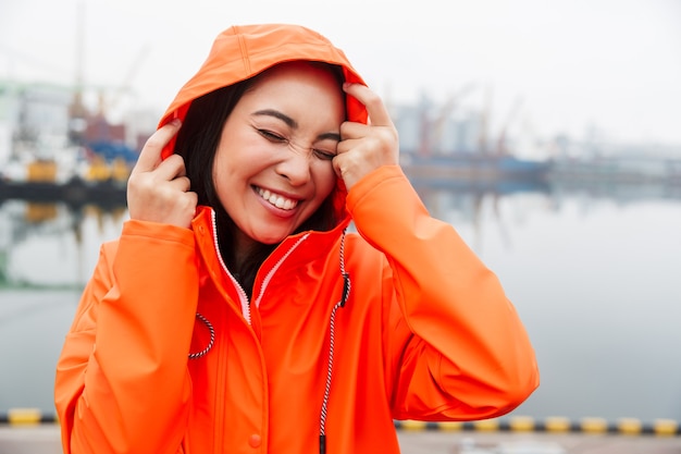 Sorridente giovane donna asiatica che indossa un impermeabile che trascorre del tempo all'aperto camminando sulla costa, posando in un cappuccio