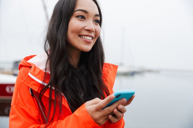 Sorridente giovane donna asiatica che indossa un impermeabile che trascorre del tempo all'aperto camminando sulla costa, usando il telefono cellulare