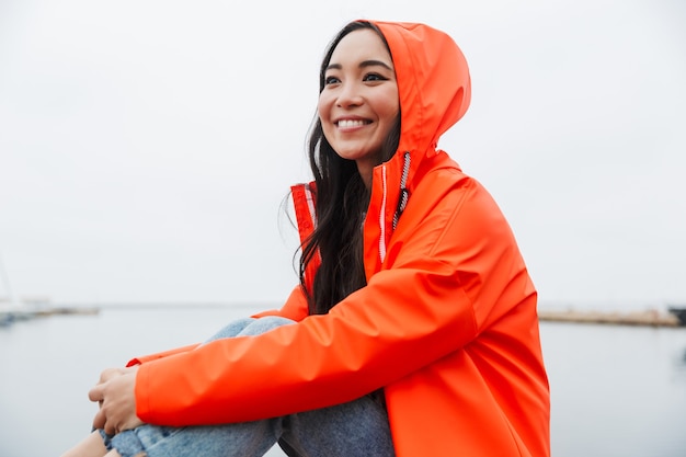 Sorridente giovane donna asiatica che indossa un impermeabile che trascorre del tempo all'aperto camminando sulla costa