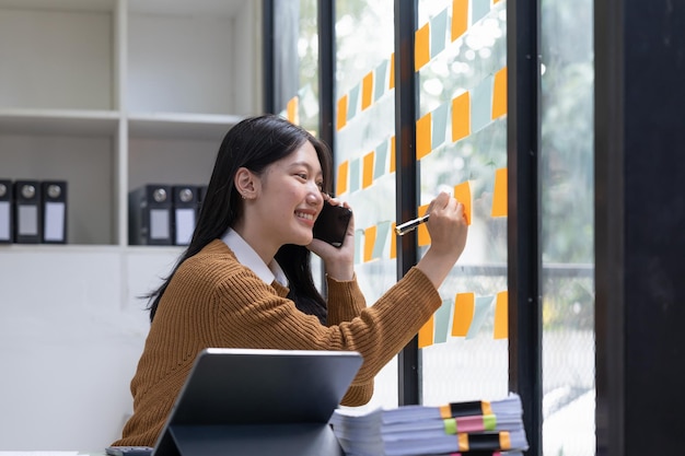 Sorridente giovane donna asiatica che scrive su foglietti adesivi sulla finestra in ufficio creativo