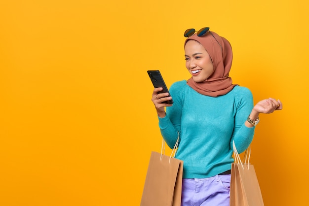 Sorridente giovane donna asiatica che utilizza un telefono cellulare e tiene in mano le borse della spesa su sfondo giallo