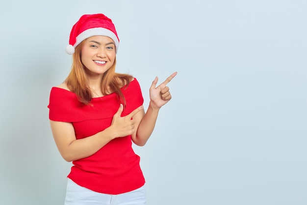 Sorridente giovane donna asiatica con cappello di Natale che punta il dito sullo spazio della copia su sfondo bianco