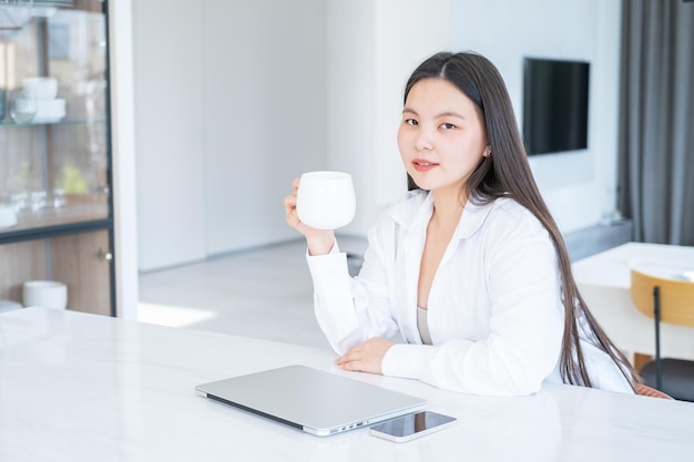 sorridente giovane donna asiatica in camicia bianca con una tazza di tè o caffè e laptop a casa moderna