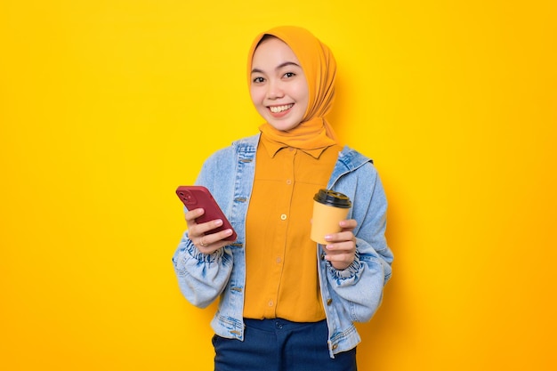 Sorridente giovane donna asiatica in giacca di jeans in possesso di telefono cellulare e tazza di caffè isolato su sfondo giallo