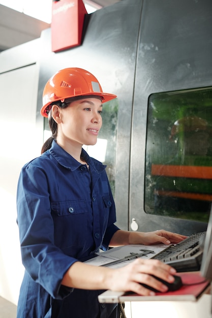 Sorridente giovane donna asiatica in hardhat operante macchina CNC durante il controllo delle impostazioni sul computer nel negozio della fabbrica